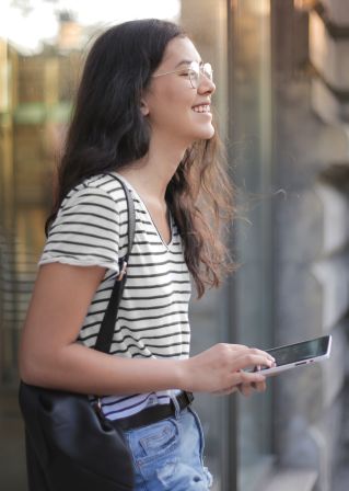 Side view of teen female in casual outfit and glasses browsing smartphone while smiling standing on street